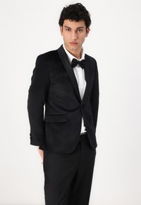 Jachetă de tuxedo neagră cu reveruri din satin, închidere cu un singur buton și buzunare frontale. Purtați peste o cămașă albă și o cravată din bumbac neagră.