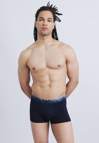 Ανδρας χωρίς μπλούζα με μακριά dreadlocks στέκεται μπροστά σε απλό φόντο φορώντας μαύρα εσώρουχα Emporio Armani με μπλε ζώνη στη μέση.