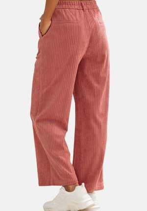 Person wearing pink wide-leg corduroy trousers with elastic waistband and white sneakers, standing with hands in pockets.