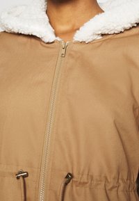 Detailní záběr na béžovou bundu s předním zipem, bílou fleecovou kapucí a nastavitelným pásem.