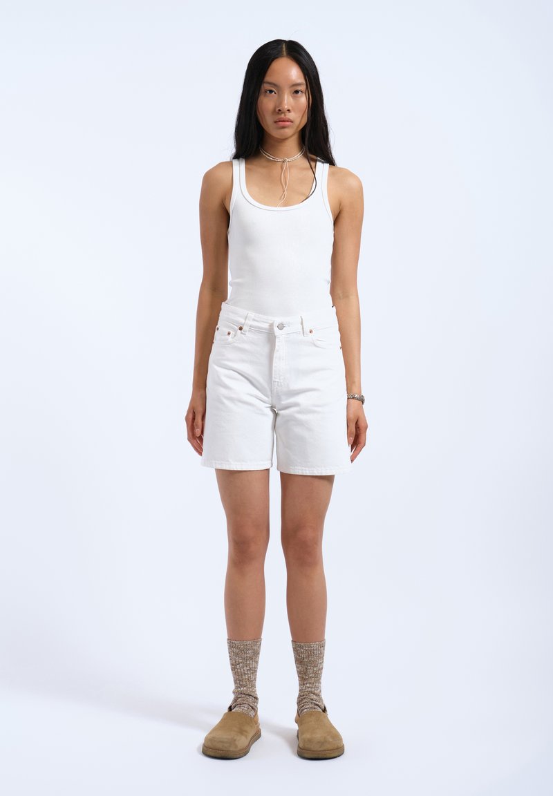 Top blanco sin mangas y pantalones cortos de mezclilla blancos, combinados con zapatos marrones y calcetas de punto beige. Diseño simple y casual, con líneas limpias.