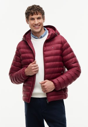 Jeune homme souriant en train de fermer une doudoune bordeaux par-dessus un pull blanc et une chemise bleue sur un fond uni.