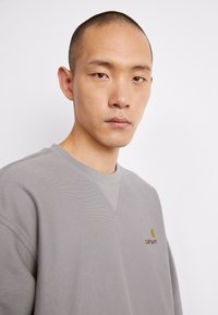 Sudadera gris con un acabado texturizado, cuello redondo y detalles de costura visibles. Presenta un pequeño logo amarillo en el pecho.