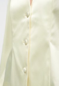 Nahaufnahme einer elfenbeinfarbenen Satinbluse mit drei bezogenen Knöpfen und sichtbaren Nähten auf glattem Stoff.