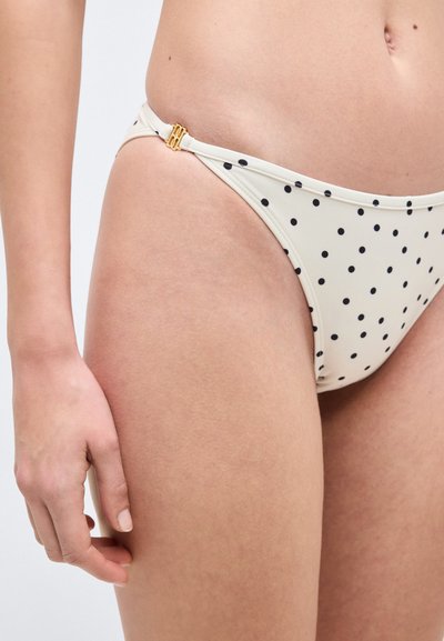 Λευκό κάτω μέρος μπικίνι με μαύρα πουά, χρυσό πλευρικό κλείσιμο, φορεμένο από ένα άτομο που δείχνει το ισχίο, το μηρό και το χέρι ενάντια σε ουδέτερο φόντο.