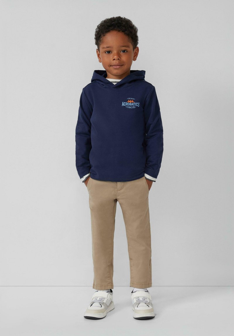 Dunkelblaue Kapuzenjacke mit einem Logo, beige Chinos und weißen Sneakers mit grauen Akzenten. Glatte Textur und lässiges Design.