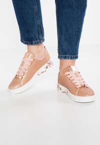 Zapatillas de purpurina en color oro rosado con cordones rosas, acentuadas por una suela blanca y diseños florales en los talones. Textura suave y forma redondeada.