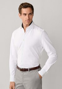 Mężczyzna w białej koszuli z długim rękawem zapinanej na guziki, szarych spodniach i brązowym skórzanym pasku, stojący z jedną ręką w kieszeni na jednolitym tle.