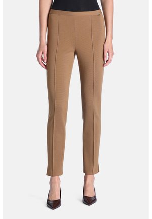 Modèle féminin portant un pantalon ajusté couleur camel avec des détails de coutures verticales et des talons aiguilles foncés à bout pointu sur fond blanc.