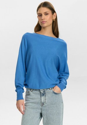 Pulover oversize albastru cu decolteu larg și mâneci lungi, fabricat din material moale; asociat cu blugi decolorati.
