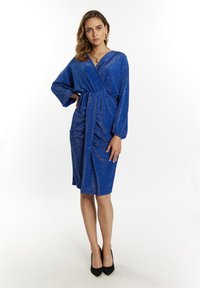 Mėlyna blizgi suknele su ilgomis rankovėmis, susiraukšlėjusia juosmens detale ir iki kelių ilgio apačia, derinama su juodomis aukštakulnėmis bateliais.