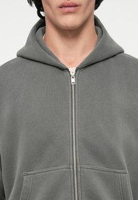 Harmaa vetoketjuhuppari, jossa on etutasku, valmistettu pehmeästä materiaalista. Varustettu nyörivedettävällä hupulla ja hopeisella vetoketjulla.