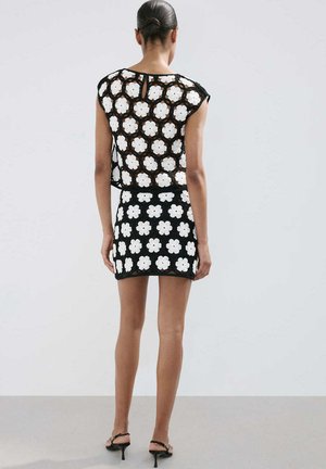 Mujer con moño que lleva un top de crochet floral en blanco y negro y una mini falda a juego, de pie de espaldas sobre un fondo liso.