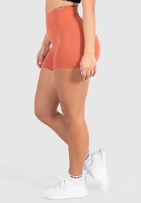 Pantaloni corti a vita alta color ruggine realizzati in tessuto elasticizzato, con una texture liscia e un design aderente. Indossati con scarpe da ginnastica bianche.