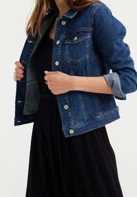 Jeansjacka i mörkblått med silverfärgade metallknappar, två bröstfickor, uppvikta ärmar och en lätt urtvättad textur, tillsammans med en svart klänning.