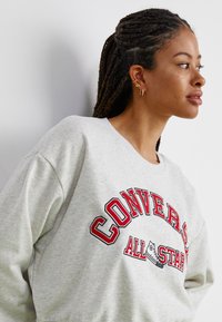 Sudadera gris claro hecha de tela suave, que presenta un bold rojo bordado con el texto "CONVERSE ALL STAR" y un clásico cuello redondo.