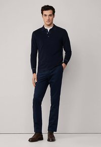 Tmavě modrá polokošile s dlouhým rukávem a límečkem, doplněná tmavě modrými chinos kalhotami. Outfit doplňují hnědé kožené boty se šněrováním. Hladká textura látky.