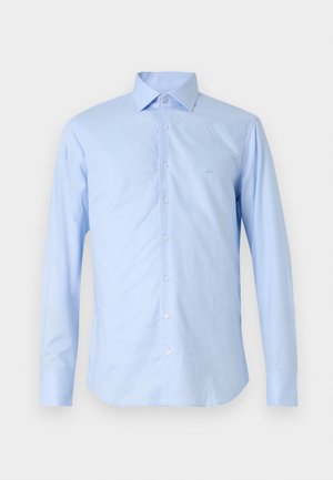 Heledad sinistea pikkade varrukatega särk, mis on valmistatud siledast kangast. Omab nööpide esit, teravat kraed ja väikest logo vasakul rinnal.