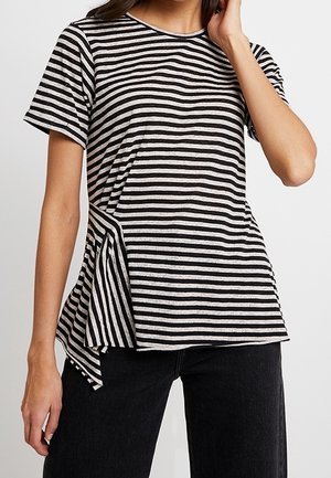 Nainen käyttää mustavalkoista vaakaraitaista lyhythihaista yläosaa, jossa on epäsymmetrinen sivuelementti, ja mustia housuja, käsi koskettaa hiuksia.