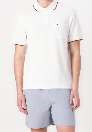 Polo blanc à manches courtes avec bordure rouge et petit logo sur la poche poitrine, associé à un short rayé bleu clair et blanc.
