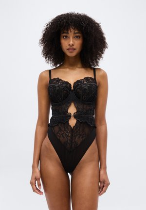 Ženska s kodrastimi lasmi, oblečena v črno čipkasto bodi z izrezi, stoji pred enostavnim svetlo sivim ozadjem.