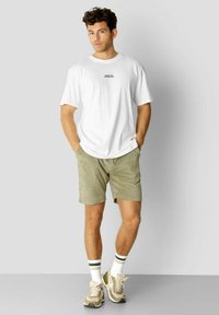 Hvit t-skjorte med korte ermer og tekst på forsiden, paired med olivenfargede shorts. Beige sporty joggesko med hvite sokker med grønne striper.