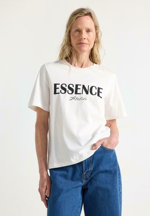 Lyshåret kvinde iført en hvid "ESSENCE Atelier" T-shirt og blå jeans, står med den ene hånd i lommen mod en ensfarvet baggrund.