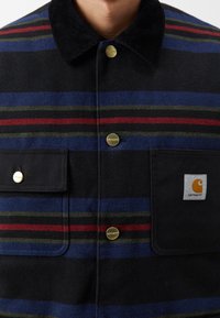 Gestreiftes Flanellhemd in Marineblau, Grün und Rot mit einem schwarzen Cordkragen, zwei vorderen Taschen und goldenen Knöpfen. Carhartt-Logo sichtbar.