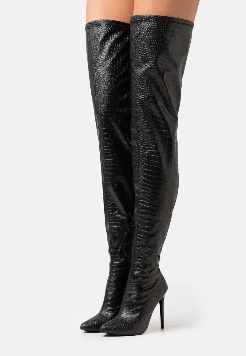 Botas negras sobre la rodilla con textura de charol, punta afilada y tacones de aguja. Presentan un cierre con cremallera en el lateral.