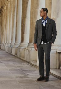 Gray tweed suit with a black vest, white shirt, and blue patterned scarf; black shoes with brown soles. Marble column backdrop.