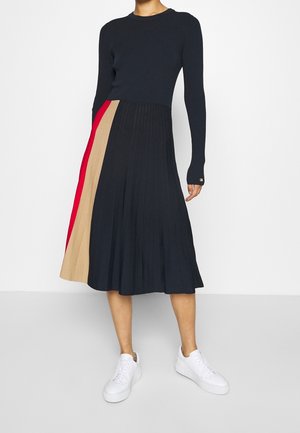 Gebreide jurk met een strak, donkerblauw bovenstuk en een geplooide rok met diagonale secties in rood, beige en navy. Witte sneakers maken de look af.