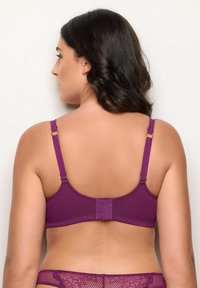 Žena s tmavými vlasy, oblečená v fialové texturované podprsence a krajkovém prádle, zobrazená zezadu na jednobarevném světlém pozadí.