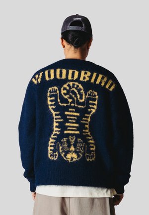 Person iført en navyblå sweater med et gult tigerdesign og "WOODBIRD" tekst på ryggen, kombineret med en sort netkasket.