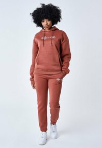 Conjunto de sudadera con capucha y pantalones jogger en color óxido, fabricado en un tejido suave, que incluye un bolsillo tipo canguro, cordones y detalles en blanco con el logo. Zapatillas blancas.
