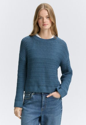Stickad tröja i en texturerad blå tyg med rund halsringning och långa ärmar, ihop med högmidjade denimjeans.