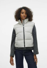 Gilet trapuntato grigio chiaro con colletto alto, zip frontale e tasche laterali. Abbinato a un maglione grigio scuro e jeans blu. Tessuto texture.