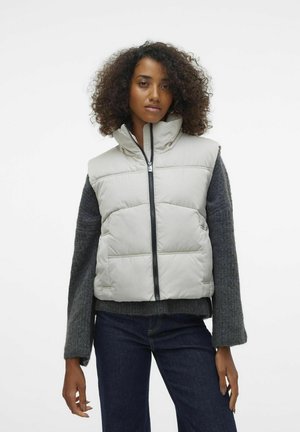Lichtgrijze puffervest met hoge kraag, ritssluiting aan de voorkant en zijzakken. Gepaard met een donkergrijze trui en blauwe spijkerbroek. Gestructureerde stof.
