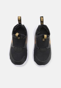 Baskets de sport noires avec tige en maille et synthétique, bande Velcro, semelle blanche et accents dorés sur la languette du talon et le logo latéral.