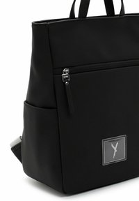 Sac à dos noir avec poche avant à fermeture éclair, poches latérales, deux poignées supérieures et un écusson comportant une lettre "Y" stylisée à l'avant.