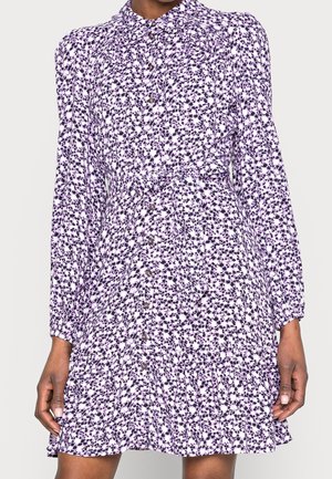 Mujer con un vestido de manga larga y botones, con un pequeño estampado floral púrpura y una cintura ceñida atada con un cinturón de tela a juego.