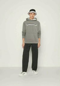 Siva kapuca z zategami in logotipom, črne hlače, bele superge in črna-bela vedro kapa. Gladek material in sproščen kroj.