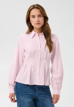 Femme aux longs cheveux bruns portant un chemisier rose clair boutonné et un jean bleu sur un fond clair uni.