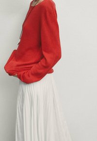 Red knitted sweater with long sleeves, paired with a flowing white pleated skirt, showcasing a smooth texture and layered appearance.