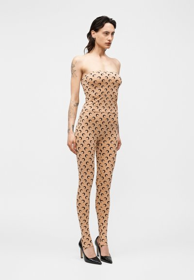 Model oblečen v bež kombinezon brez naramnic s črnimi vzorci polmeseca in črnimi čevlji z visokimi petami, stoji na enobarvni podlagi.