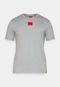 Camiseta de manga corta de color gris claro con cuello redondo y un parche rectangular rojo etiquetado "HUGO" centrado en el pecho.