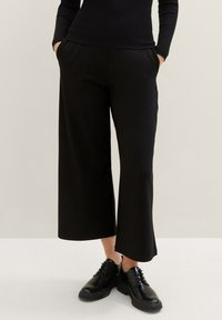 Pantaloni neri a gamba larga con texture liscia, dotati di tasche laterali e vita aderente, abbinati a scarpe nere con lacci.