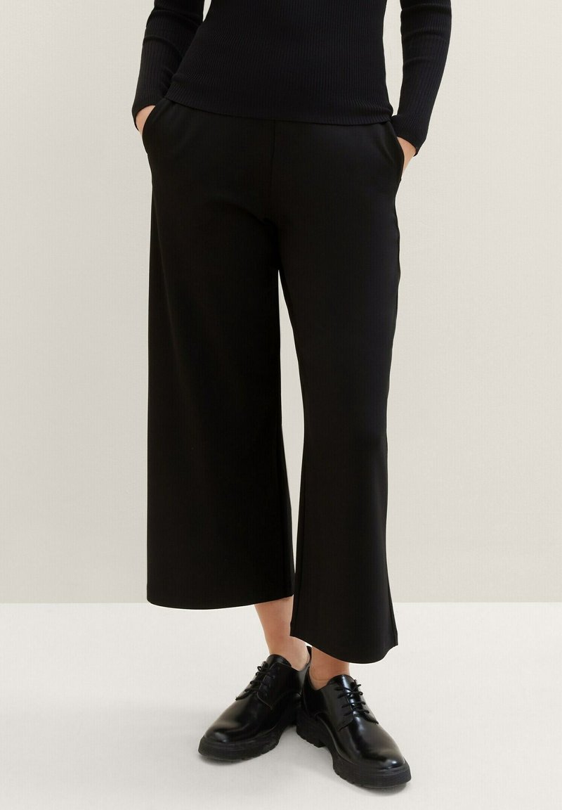 Pantaloni neri a gamba larga con texture liscia, dotati di tasche laterali e vita aderente, abbinati a scarpe nere con lacci.