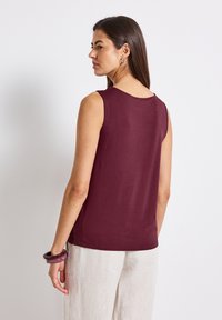 Burgundyfärgad ärmlös topp med rund halsringning, tillverkad av mjukt tyg. Bärs tillsammans med beige linbyxor som visar en slät textur och enkel design.