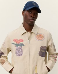 Μπεζ καμβάς μπουφάν με διάφορα πολύχρωμα γραφικά σχέδια, που περιλαμβάνουν λουλούδια, ένα καπέλο, φαγητό και ένα κερί. Διαθέτει φερμουάρ και πλευρικές τσέπες.