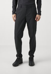 Black tapered trousers made from smooth, lightweight fabric. Features a belt loop waistband, front pockets, and ankle cuffs. Paired with black sneakers.